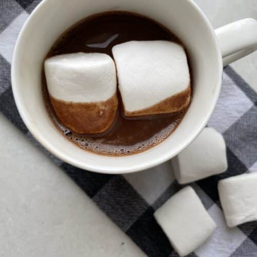 Low sodium hot chocolate , topped with two large marshmallows, on a black and white tea towel surrounded by more large marshmallows.