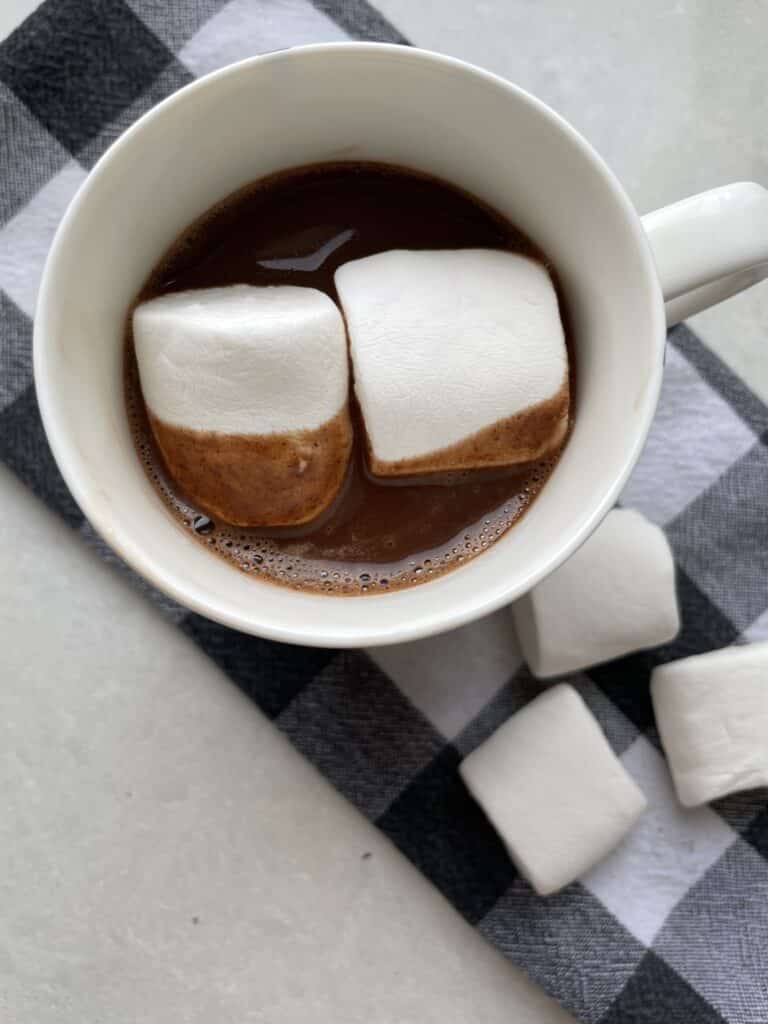 Low sodium hot chocolate , topped with two large marshmallows, on a black and white tea towel surrounded by more large marshmallows.
