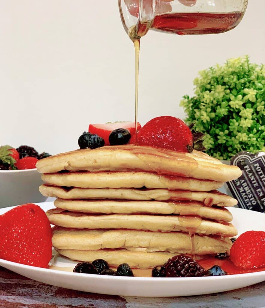 Maple syrup being poured over stack of pancakes topped with berries