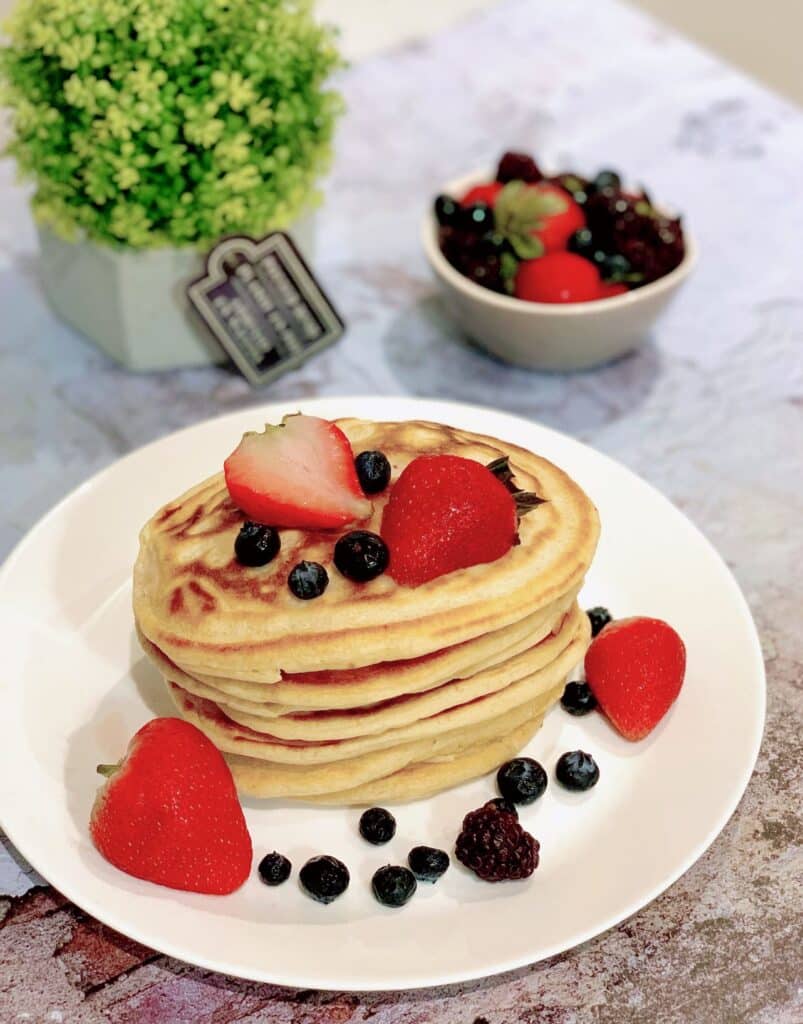 A stack of pancakes on white plate topped with berries and. Green plant and berries in background