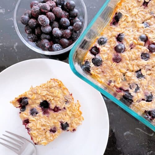 Protein baked oats in a baking dish with a side of frozen blueberries and a piece of baked oatmeal on a white dish with a fork