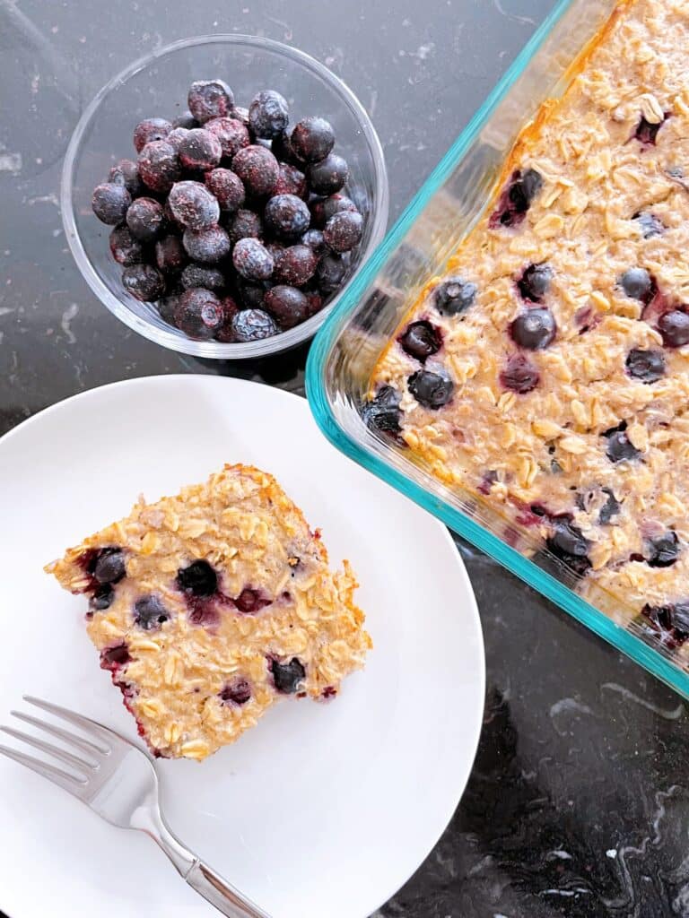 Protein baked oats in a baking dish with a side of frozen blueberries and a piece of baked oatmeal on a white dish with a fork
