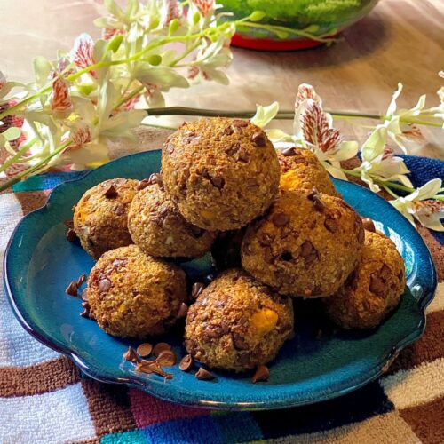 Picture of rolled pumpkin protein balls on a blue plate, on a brown checkered tea towel surrounded by white flowers and a bowl in the background