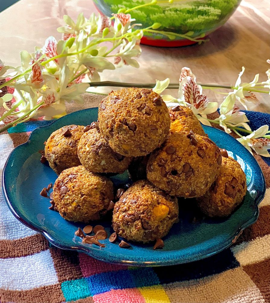 Picture of rolled pumpkin protein balls on a blue plate, on a brown checkered tea towel surrounded by white flowers and a bowl in the background 