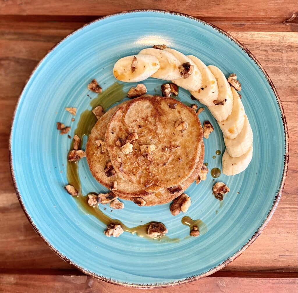 pancake on blue plate on a wood table. On the plate is a side of sliced banana and the pancakes are topped with nuts and maple syrup