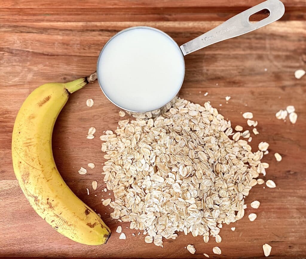 A whole banana, a cup of rolled oats and 1 cup of milk on a wood table.