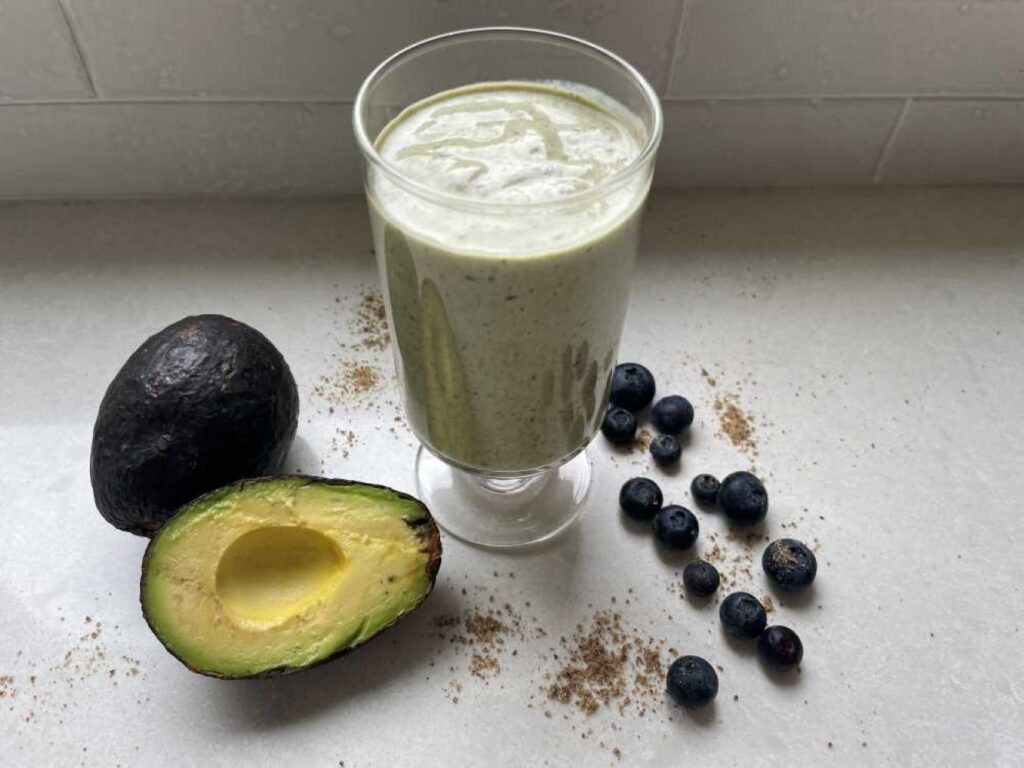 A green smoothie to lower cholesterol featured in clear glass surrounded by a cut avocado, scattered blueberries and ground flaxseed