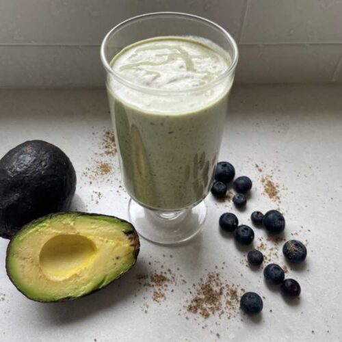 A green smoothie to lower cholesterol featured in clear glass surrounded by a cut avocado, scattered blueberries and ground flaxseed