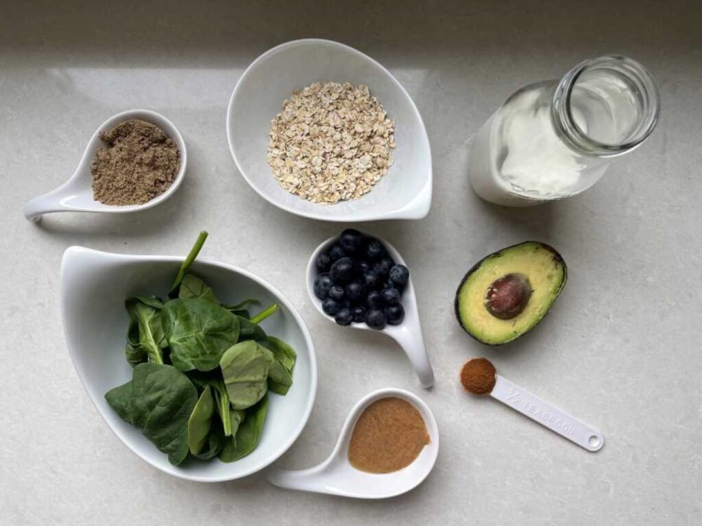 Ingredients in a smoothie to lower cholesterol displayed: ground flaxseed, fresh spinach, rolled oats, blueberries, almond butter, cinnamon, half an avocado and soy milk
