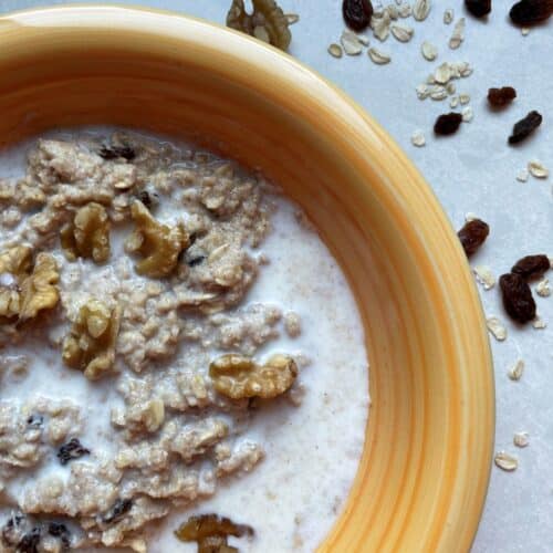 Microwave Egg White Oatmeal in a yellow bowl surrounded by oats, raisin and walnuts on a white countertop