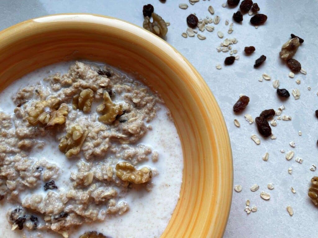 Microwave Egg White Oatmeal in a yellow bowl surrounded by oats, raisin and walnuts on a white countertop