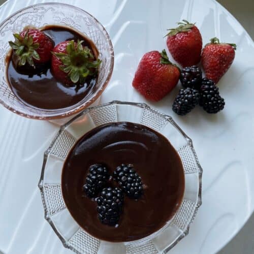 2 Oat milk chocolate pudding presented in glass jars. One with blueberries, and the other with strawberries on a white table with a side of berries.