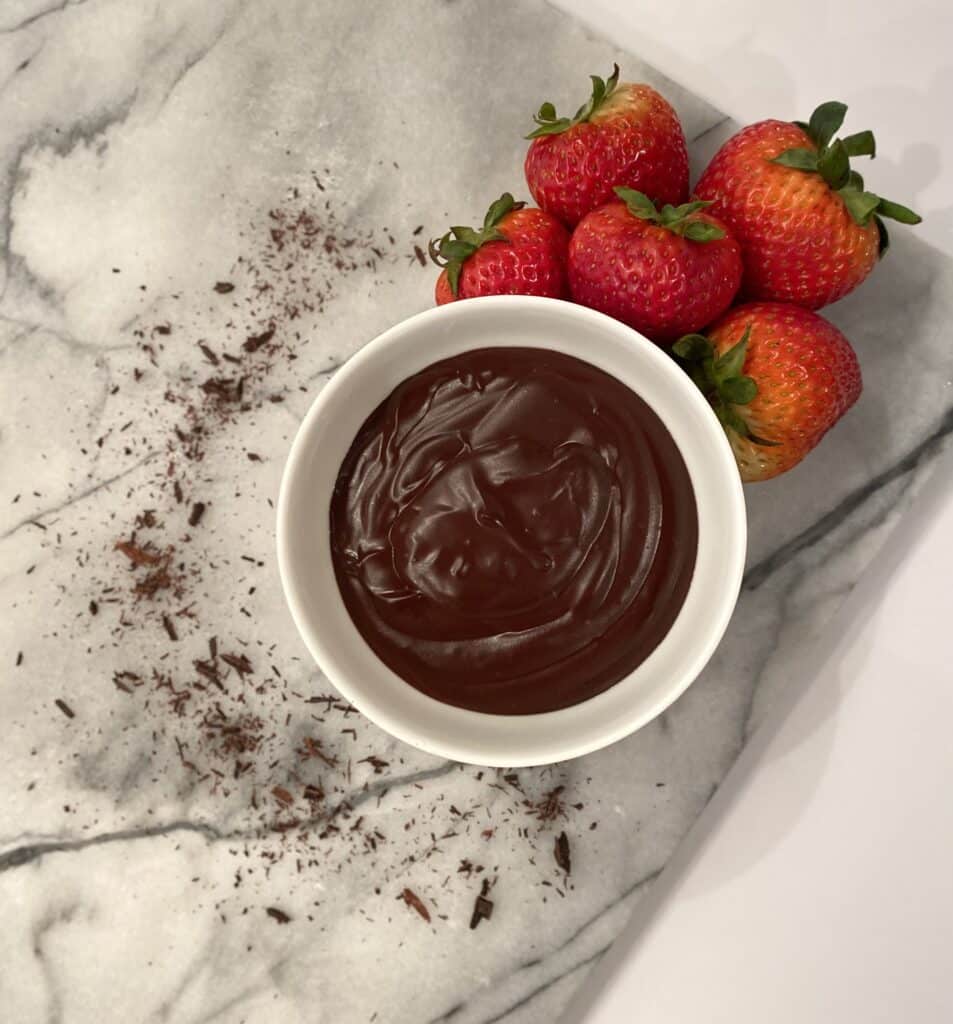 oat milk pudding in a white bowl surrounded by fresh strawberries, cocoa powder on a marble cutting board