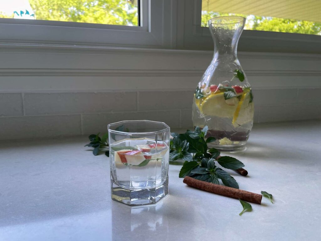 Jar of pineapple cucumber ginger lemon apple water in the background and a glass of water with some diced apples in the foreground on a countertop in front of a window