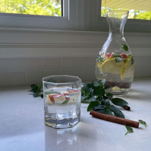 Jar of pineapple cucumber ginger lemon apple water in the background and a glass of water with some diced apples in the foreground on a countertop in front of a window