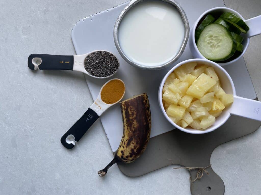 Ingredients in a pineapple smoothie for weight loss on cutting board, chia seeds and turmeric, banana in the peel, diced pineapple in measuring cup, milk in measuring cup, sliced cucumber in measuring cup