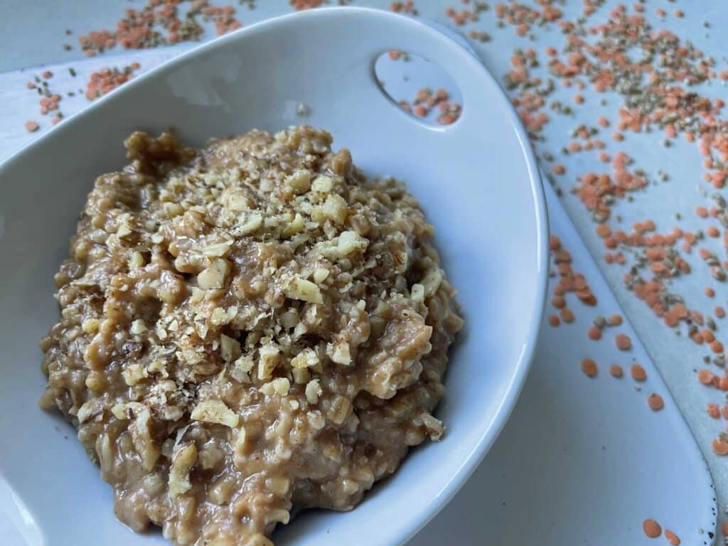 An image of vegan protein oatmeal in a white bowl topped with walnuts, and lentils around the dish.