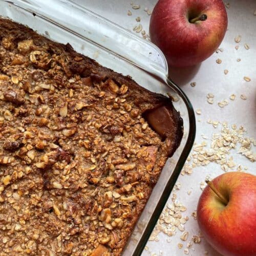 Baked oats in clear baking dish surrounded by 3 red apples and a sprinkle of oats on a white countertop.