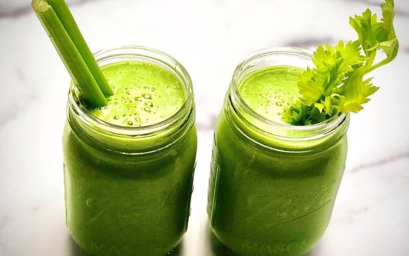 Two green smoothies in a clear glass with celery as a straw on a white background.