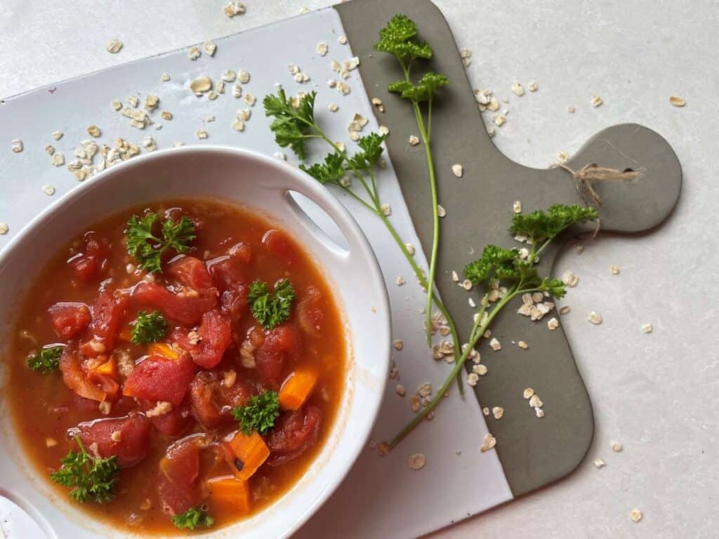 Bowl of tomato oatmeal soup in a white bowl garnished with parsley on a white and grey cutting board surrounded by a spring of parsley and a sprinkle of oats