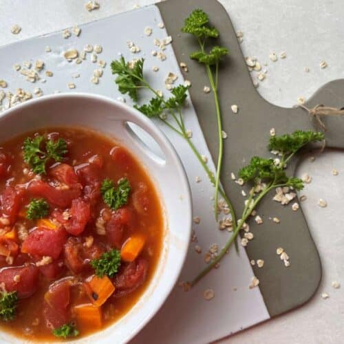 Bowl of tomato oatmeal soup in a white bowl garnished with parsley on a white and grey cutting board surrounded by a spring of parsley and a sprinkle of oats