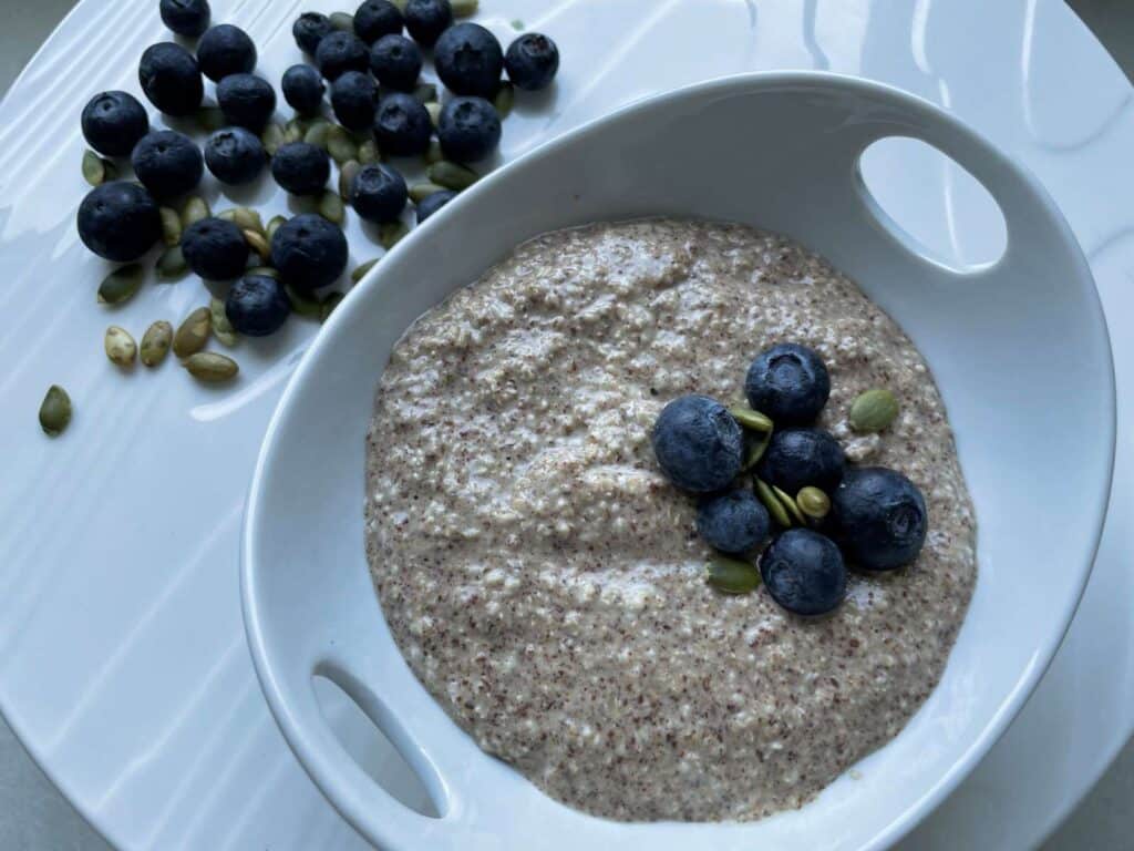 Overnight Oat bran in a white bowl topped with blueberries and pumpkin seeds surrounded by a handful of blueberries and pumpkin seeds on a white platter