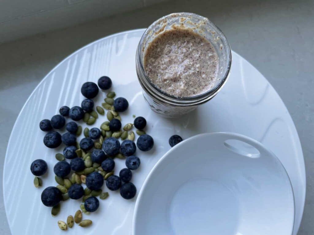 Overnight Oat bran in mason jar beside an empty white bowl and a handful of blueberries and pumpkin seeds on a white platter