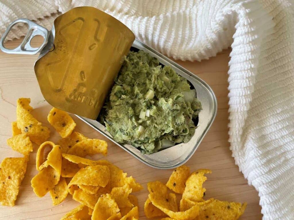 Sardine dip in sardine can surrounded by corn chips and a white towel on a wood cutting board