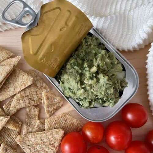Sardine dip in sardine can surrounded by cherry tomatoes, whole grain crackers and a white towel on a wood cutting board.