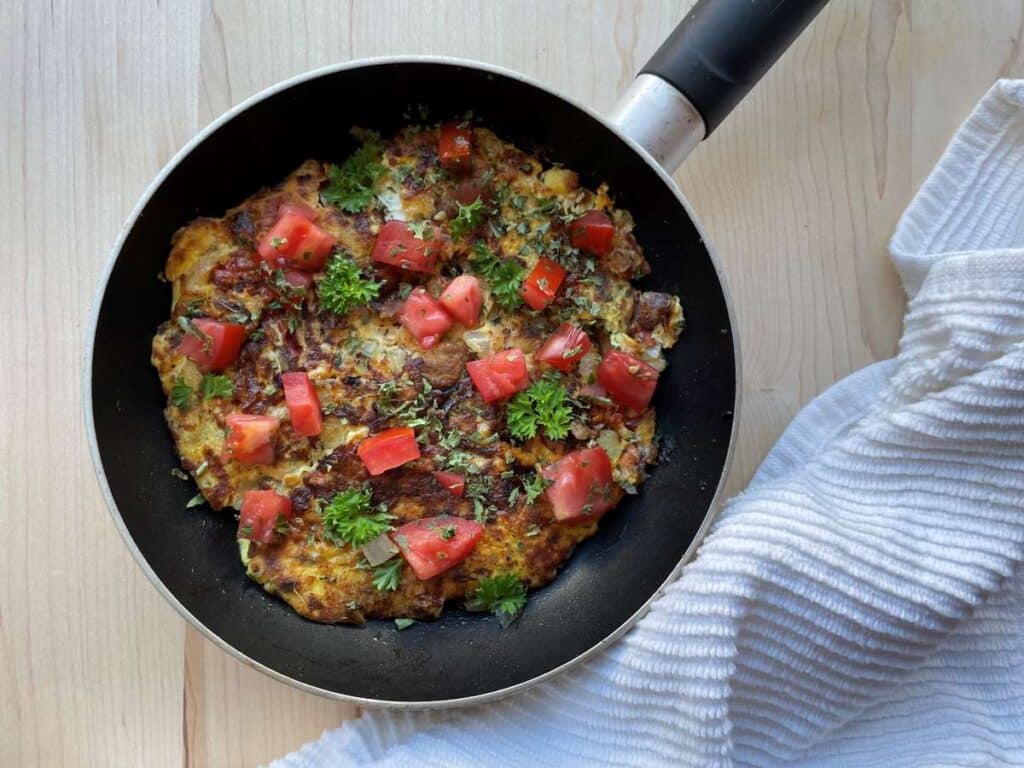 Sardine omelet in frying pan topped with tomatoes and parsley on a wood cutting board beside a white towel