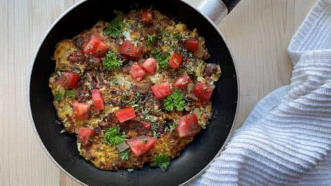 Sardine omelet in frying pan topped with tomatoes and parsley on a wood cutting board beside a white towel
