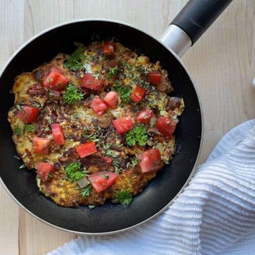 Sardine omelet in frying pan topped with tomatoes and parsley on a wood cutting board beside a white towel