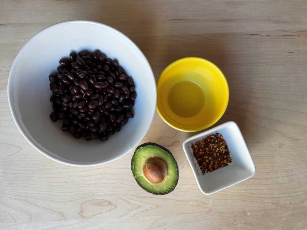Ingredients for black bean toast include black beans in white bowl, oil in yellow bowl, half of an avocado with the pit and 1 tbsp red chili peppers on a wood cutting board