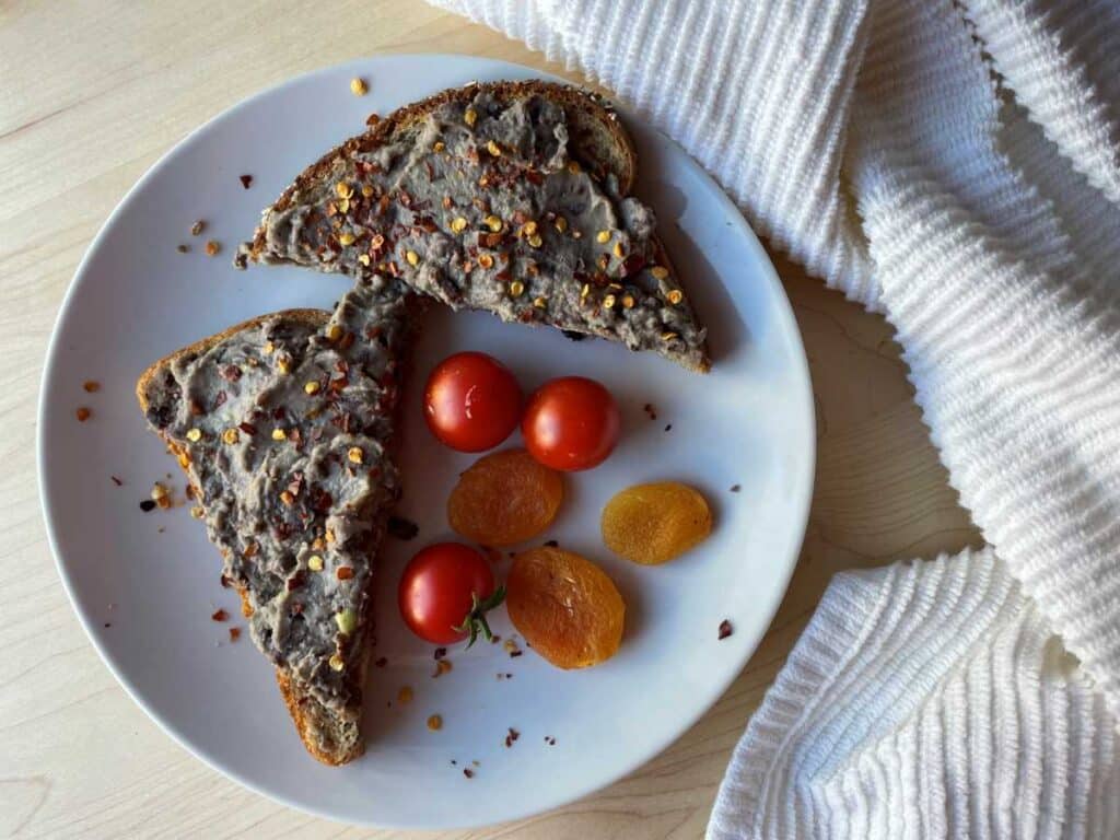 A slice of toast with black bean mixture, sliced in triangles. Garnished with red chili peppers and a side of cherry tomatoes and apricots on a white plate, surrounded by a white towel on a wood cutting board.