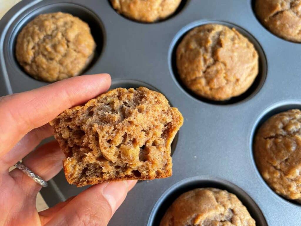 A persons hand holding a broken muffin (to show fluffy muffin consistency) between two fingers with 4 muffins in muffin tin in the background.