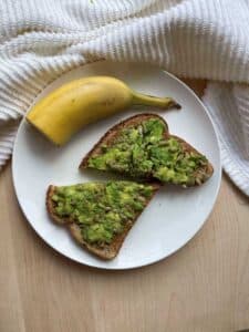 Peanut butter avocado toast and half of a banana still in it's peel on a white plate on top of a wood cutting board.