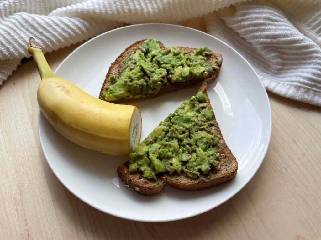 Peanut butter avocado toast and half of a banana still in it's peel on a white plate on top of a wood cutting board.