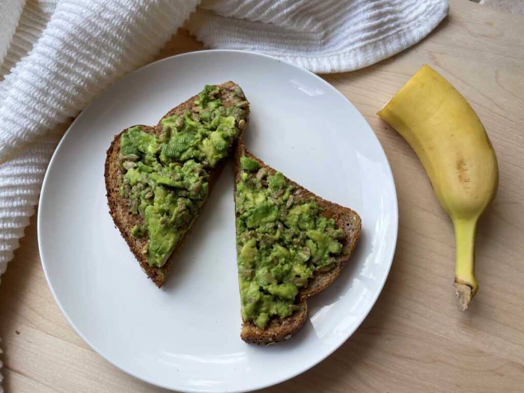 Peanut butter avocado toast on white plate with half of a banana still in it's peel beside it on a wood cutting board