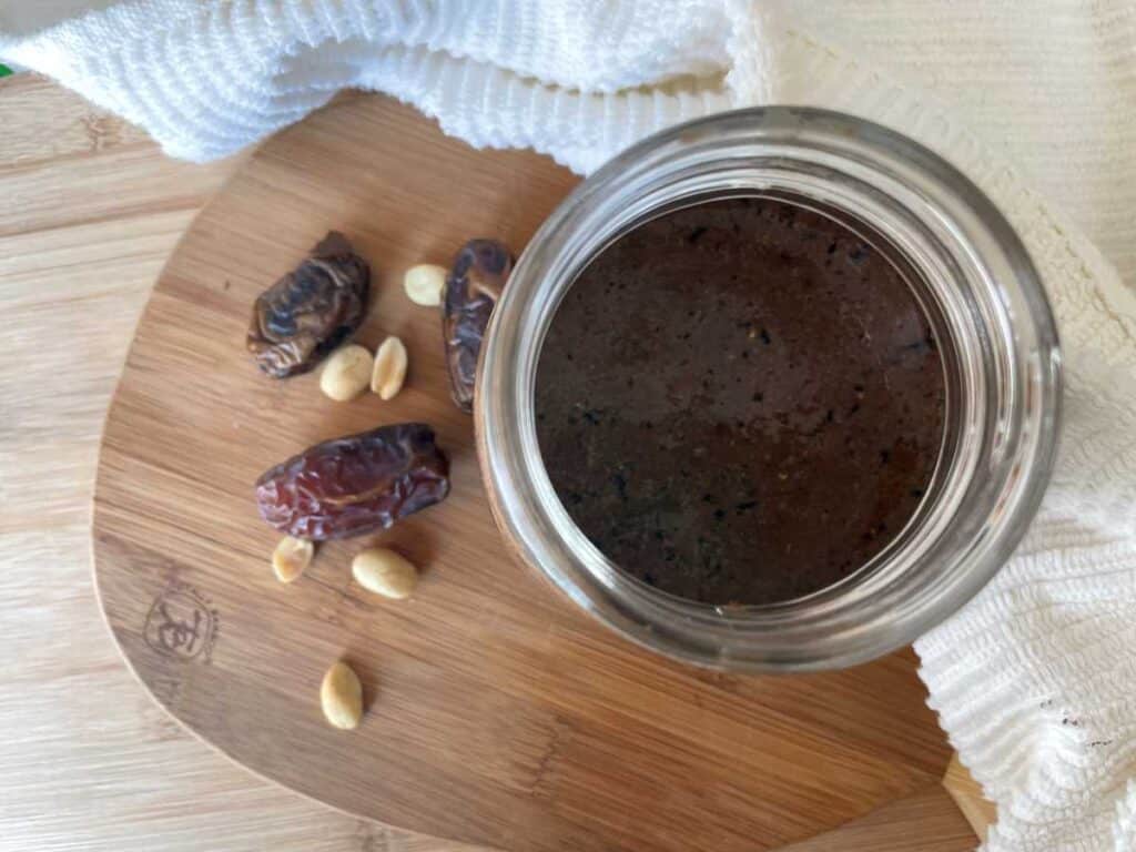 brown smoothie in mason jar on two wood cutting boards surrounded by a white towel beside dates and peanuts.