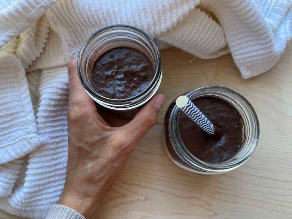 Two black bean smoothies in mason jar on a wood cutting board surrounded by a white towel. One smoothie has a blue and white straw, and the other is being held by a hand.