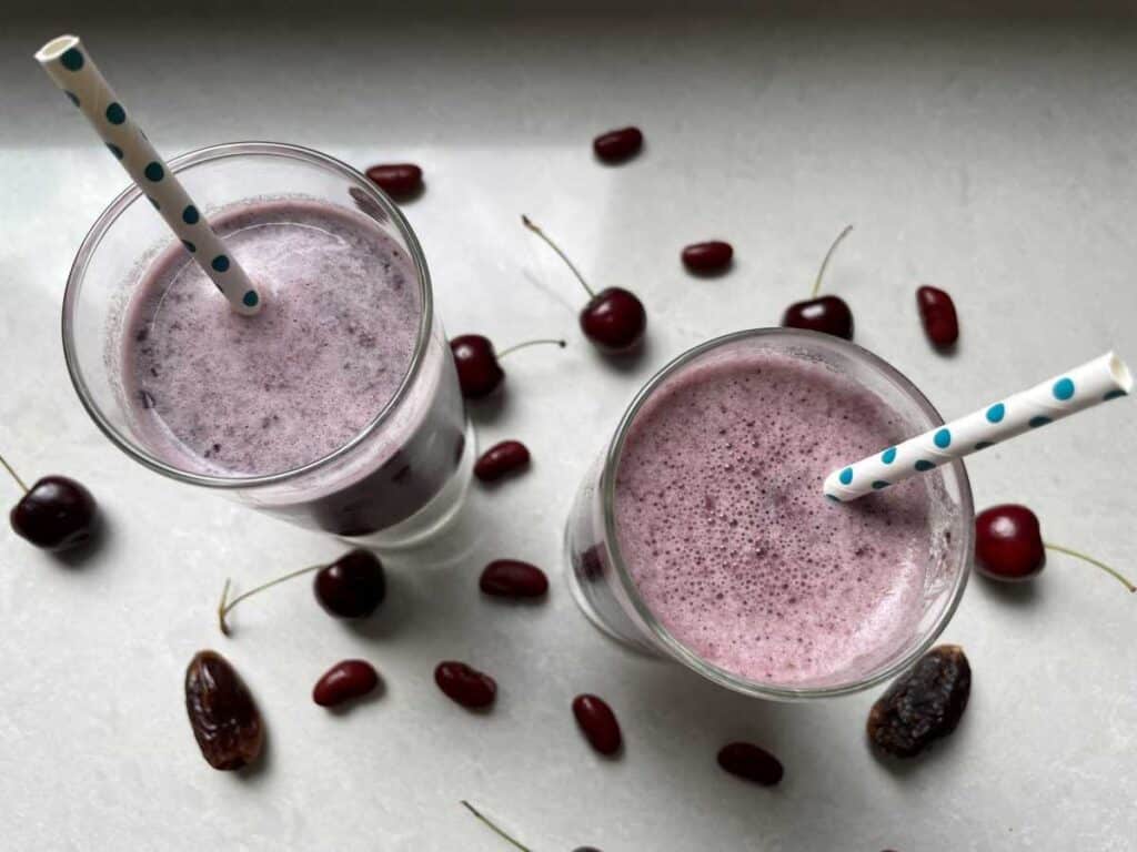 Purple smoothies in glass cups with blue straws surrounded by cherries, red kidney beans and dates on a white countertop.