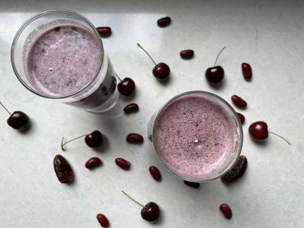 Two red bean smoothies, surrounded by cherries, red kidney beans and dates on a white countertop.