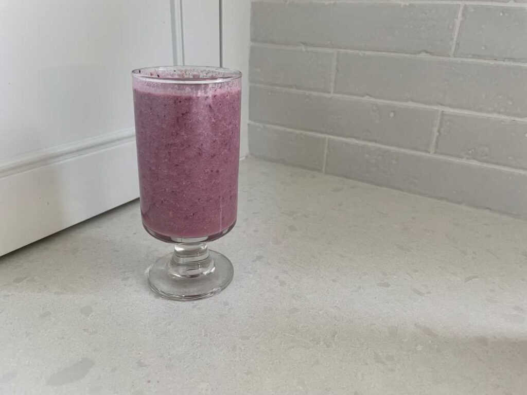 Purple smoothie in clear glass on white counter between white cupboard and white backsplash.