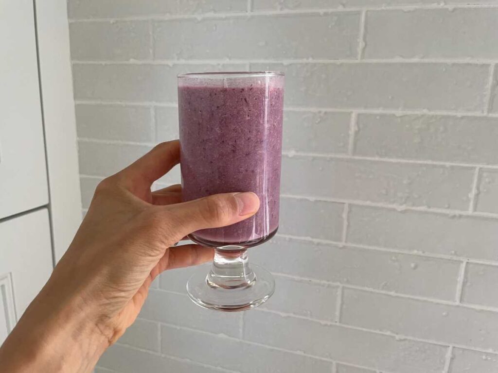 Purple smoothie in clear glass being held by a hand in front of white backsplash.