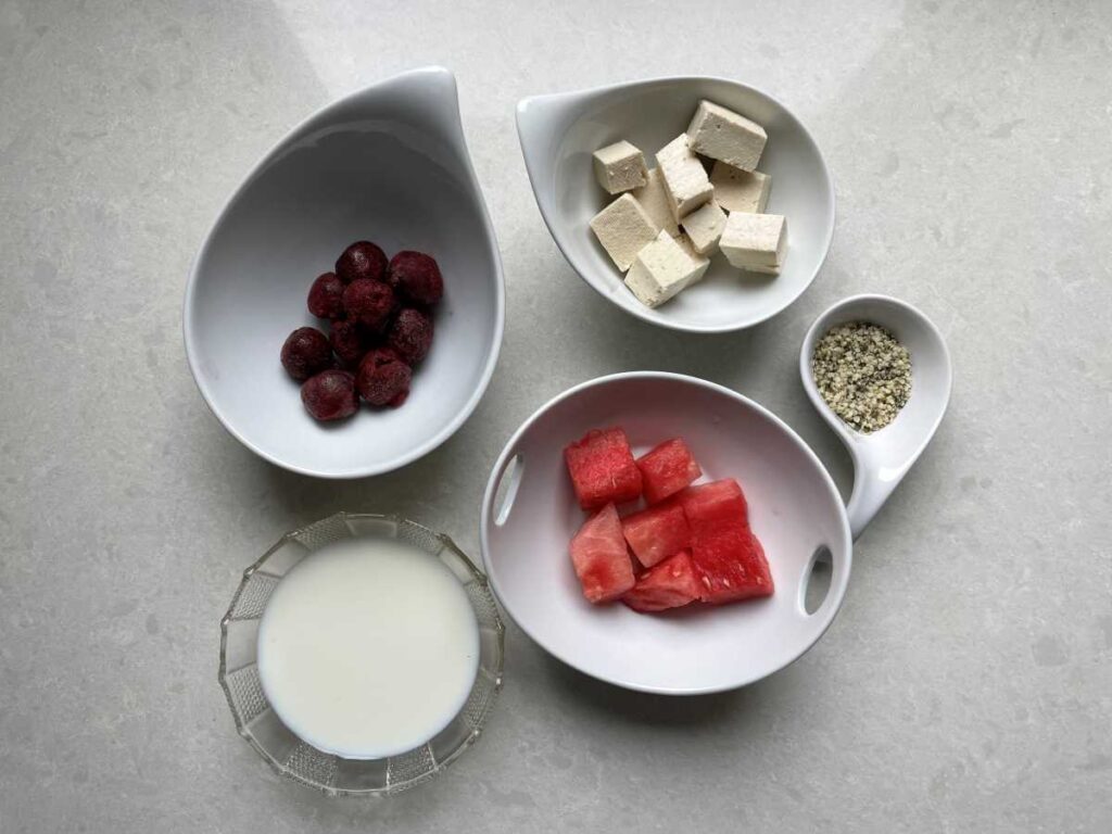 Ingredients displayed in white bowl on white counter include frozen cherries, cubed white tofu, hemp hearts, cubed watermelon and milk.