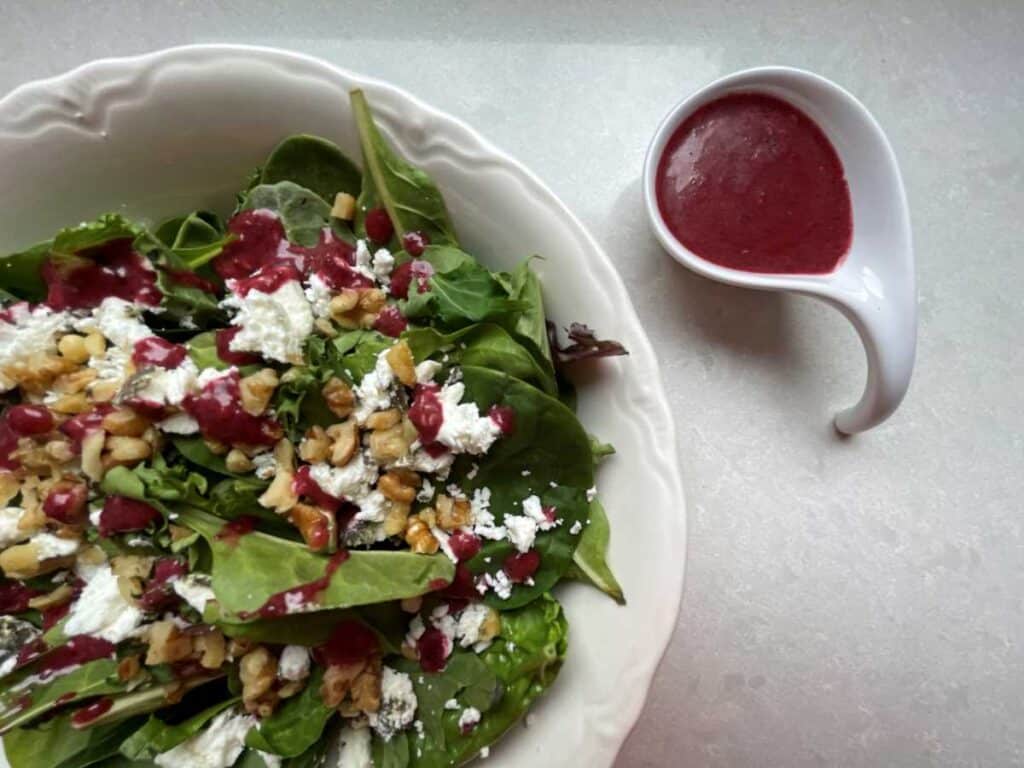 Salad in white bowl topped with goat cheese, walnuts and red Cherry Vinaigrette. Beside salad bowl is a small portion of Cherry Vinaigrette in a small white pourable bowl.