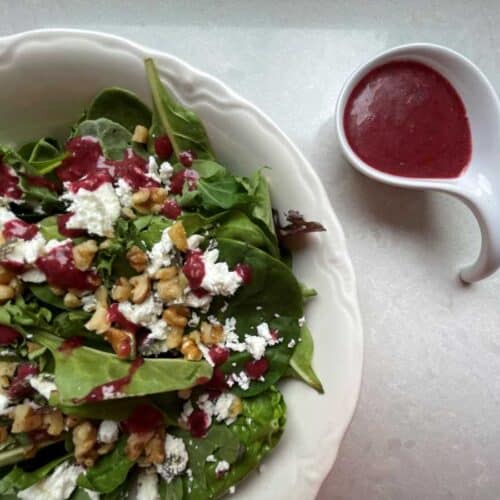 Salad in white bowl topped with goat cheese, walnuts and red Cherry Vinaigrette. Beside salad bowl is a small portion of Cherry Vinaigrette in a small white pourable bowl.