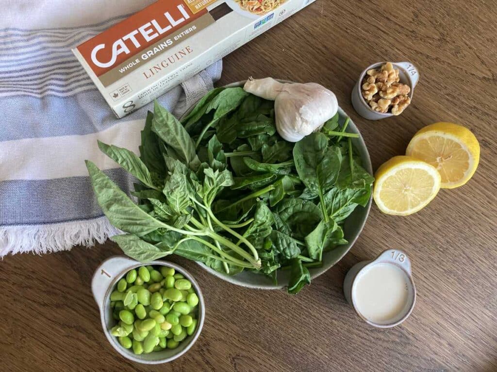 Ingredients for Edamame Pasta Sauce presented on wood table beside  a white and blue tea towel. Ingredients include a box of spaghetti noodles, a bowl of spinach and basil with garlic. A sliced lemon. Soy milk, walnuts and edamame.