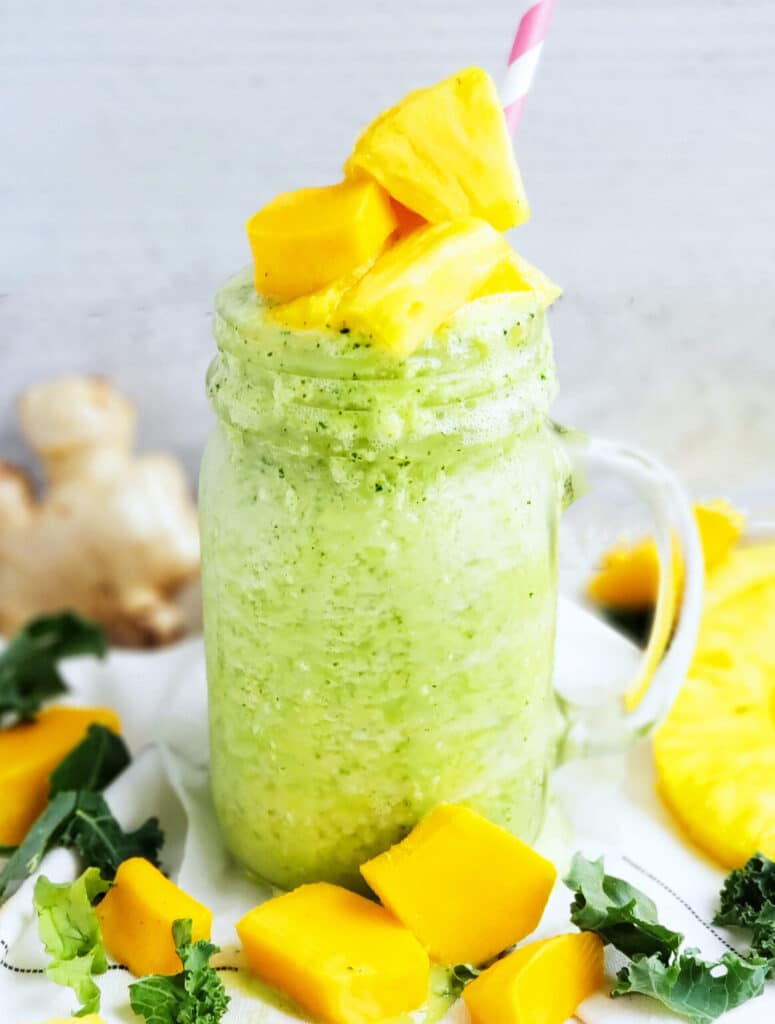 Green smoothie in a mason jar with a handle, topped with mango cubes and a red and white straw surrounded by more cubes of mango.