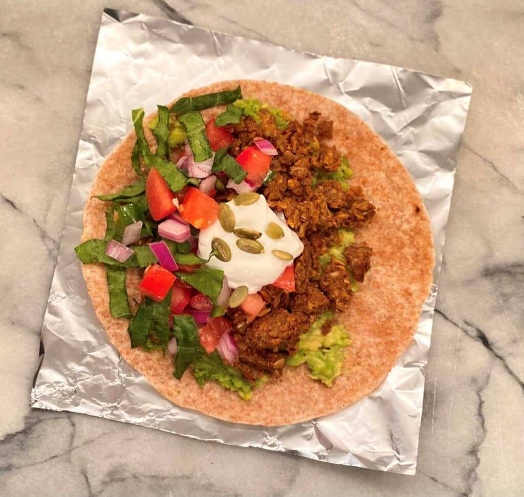 An open wrap on tin foil that has lentil burrito filling, homemade salsa, lettuce, avocado and topped with Greek yogurt.